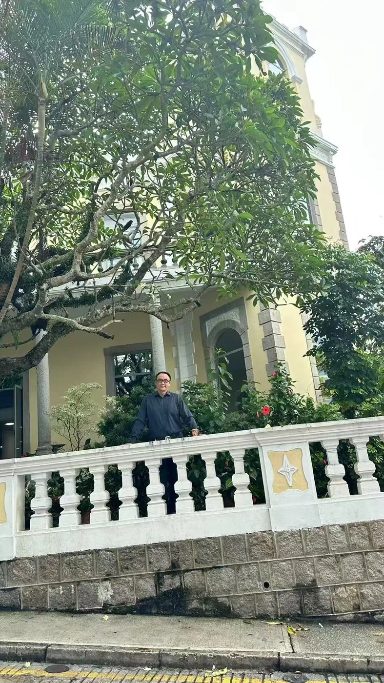 Sean Own standing on the balcony of the Alves de Mesquita colonial mansion in Macao, in front of a tree and a yellow facade with white balustrades