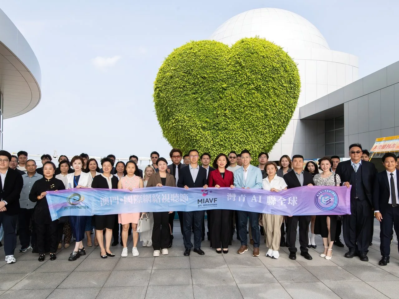Group photo of 25+ Bay Youth AI delegates outside the Macao Science Centre in front of its heart-shaped green wall, holding the MIAVF banner, 2 June 2026