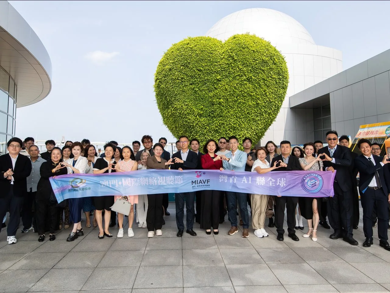 Group photo of 25+ Bay Youth AI delegates forming hand-hearts outside the Macao Science Centre in front of its heart-shaped green wall, 2 June 2026
